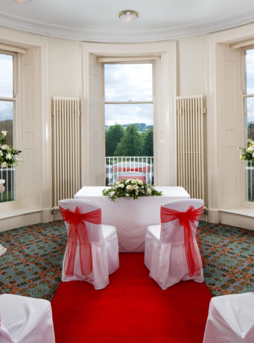 Table in a large bay window dressed in white linen and red ribbons ready for a wedding ceremony at Mercure Gloucester Bowden Hall Hotel